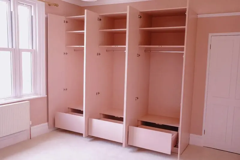 Pink painted fitted wardrobe interiors with hanging rails, shelves and pull-out drawers along a bedroom wall